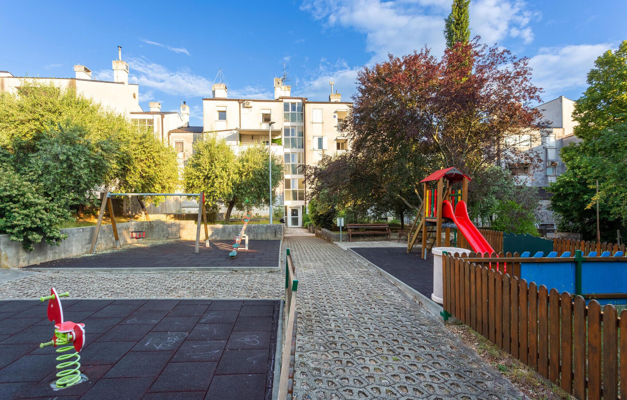 A playground with a slide and swings, surrounded by a wooden fence and trees.