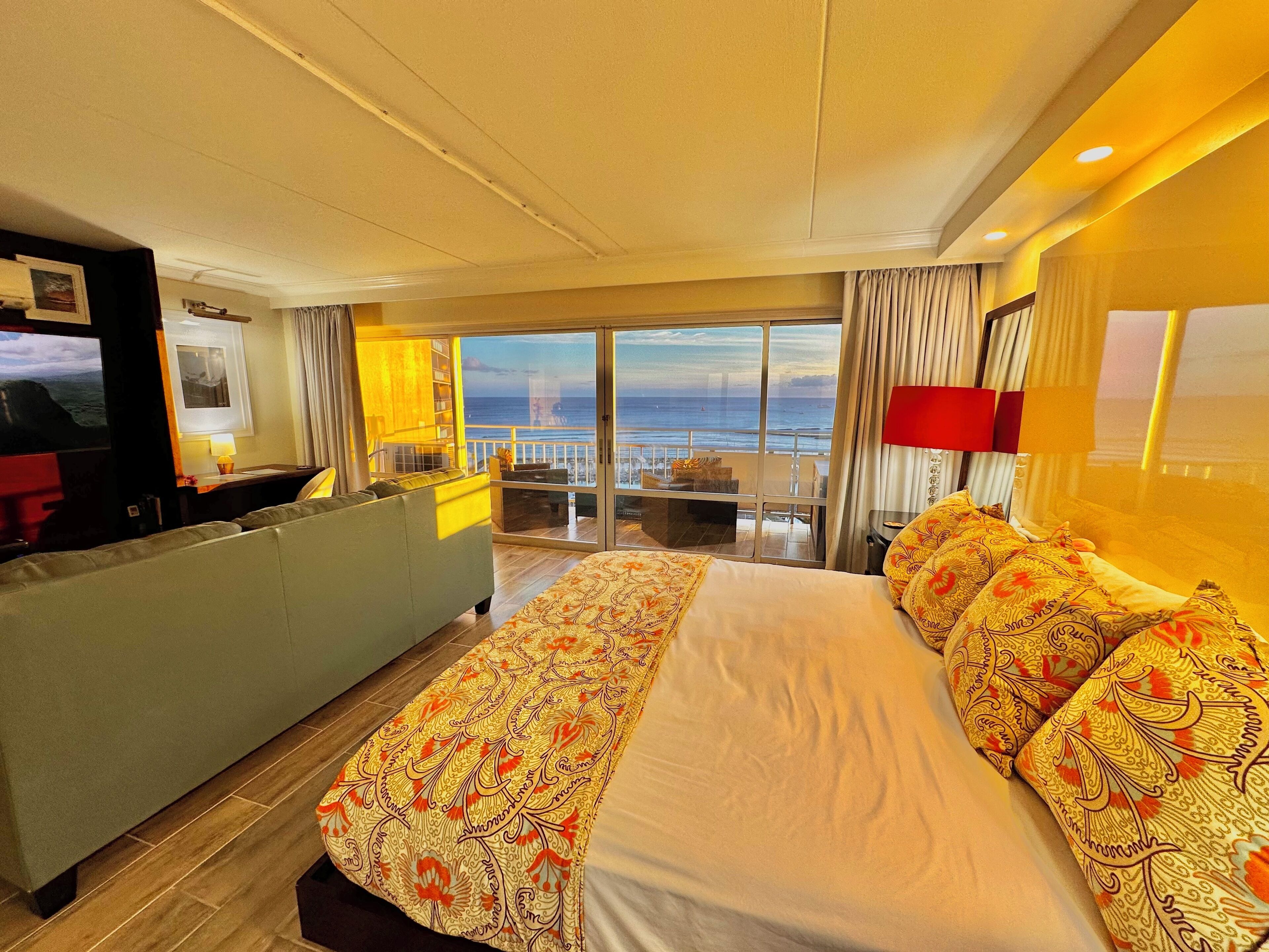 A hotel room with a large bed, a sofa, a TV, and a balcony with a view of the sea.