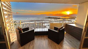 En balkon med to sorte fletsofaer, et lille bord og et skab. Balkonen har udsigt over en havn med både og en solnedgang over havet.