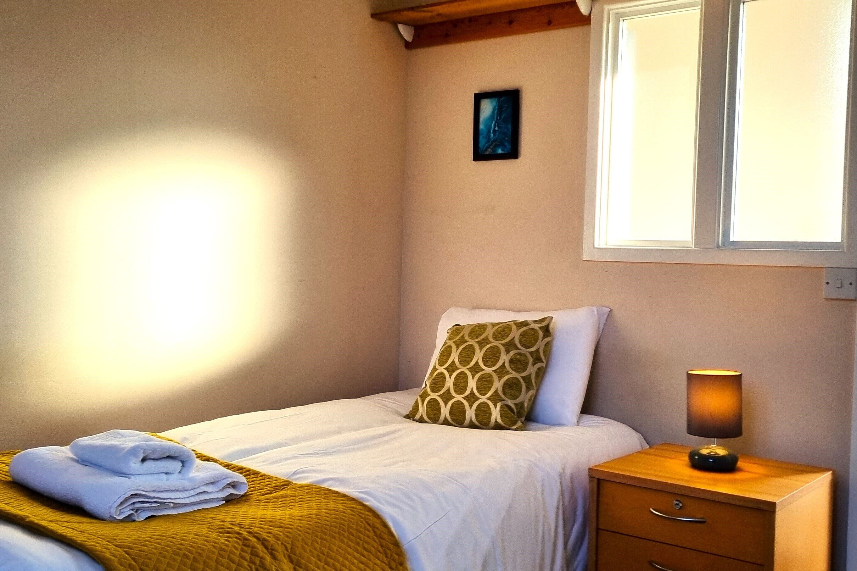 A neatly made bed with a folded towel, a patterned pillow, and a bedside lamp on a wooden nightstand.