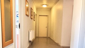 Un pasillo estrecho y bien iluminado con un radiador en la pared, una puerta al final y cuadros en la pared izquierda.