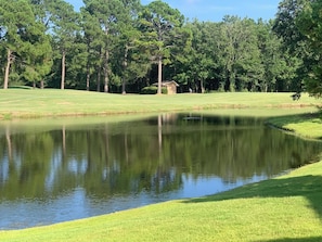Un campo de golf tranquilo con un estanque, rodeado de árboles verdes y un edificio pequeño a lo lejos.