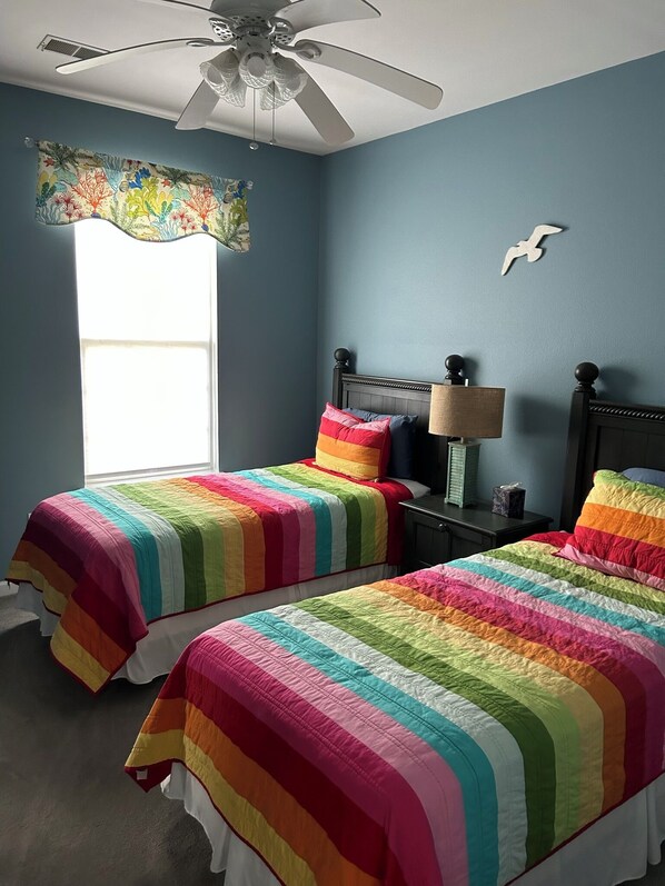 A bedroom with two beds, a ceiling fan, a nightstand, a lamp, and a window with a curtain.