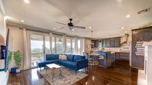 Amplia sala de estar con un sofá seccional azul, una mesa de centro y una mesa de comedor con sillas. Se observa una cocina con armarios de madera oscura y una isla grande. Se puede ver un ventilador de techo, y el espacio está bien iluminado con luz natural que entra por ventanas amplias.