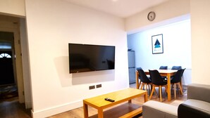Un salón con televisión de pantalla plana montada en la pared, una mesa de centro de madera, un sofá gris y una zona de comedor con mesa de madera y sillas negras.
