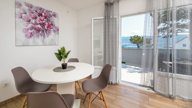 A modern dining area with a round table, four chairs, a potted plant, and a large floral painting on the wall. - Holiday home with terrace near the sea (Šilo)
