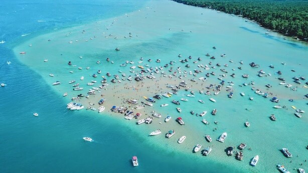 Vista aérea de uma praia com vários barcos ancorados em águas rasas, rodeada por mata densa.