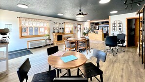 A spacious room with a fireplace, wooden flooring, and a ceiling fan. There are tables and chairs arranged for dining or meetings. - Hansen Inn and Cabins (Wall)