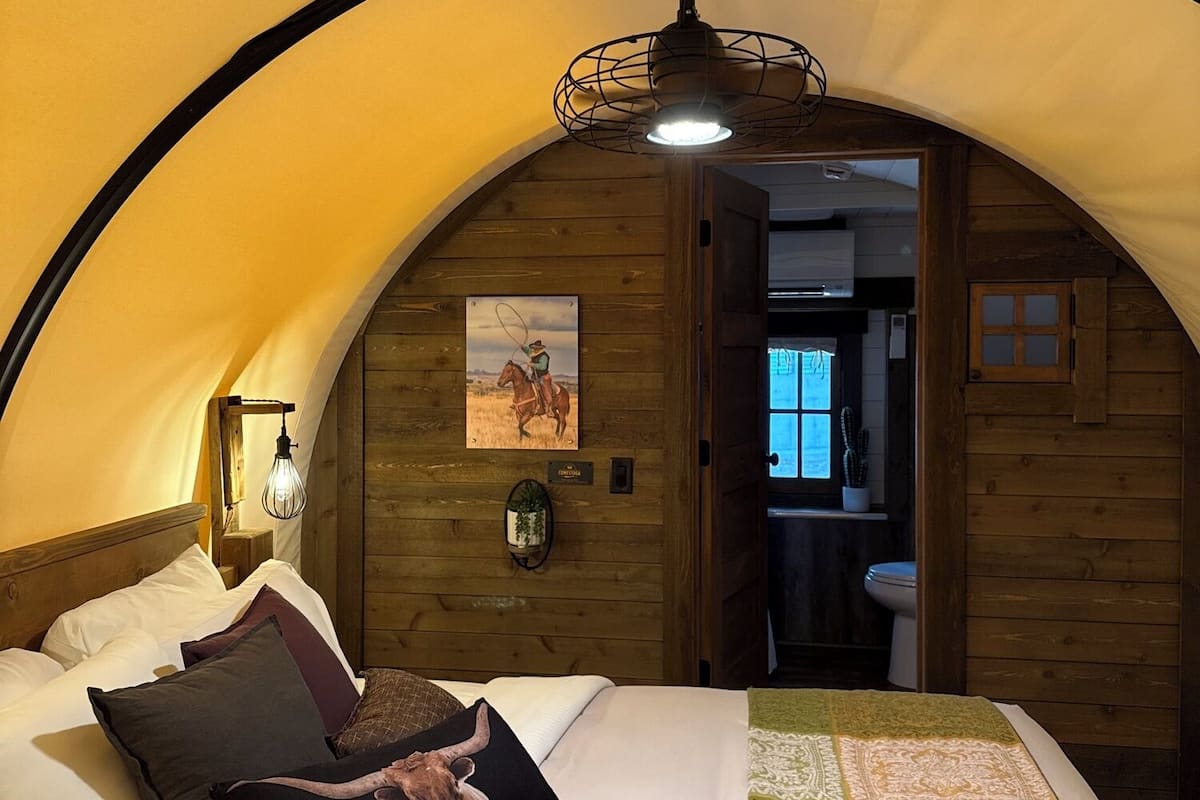 A cozy, arched bedroom with a bed, pillows, a quilt, a wall-mounted light, and a painting.