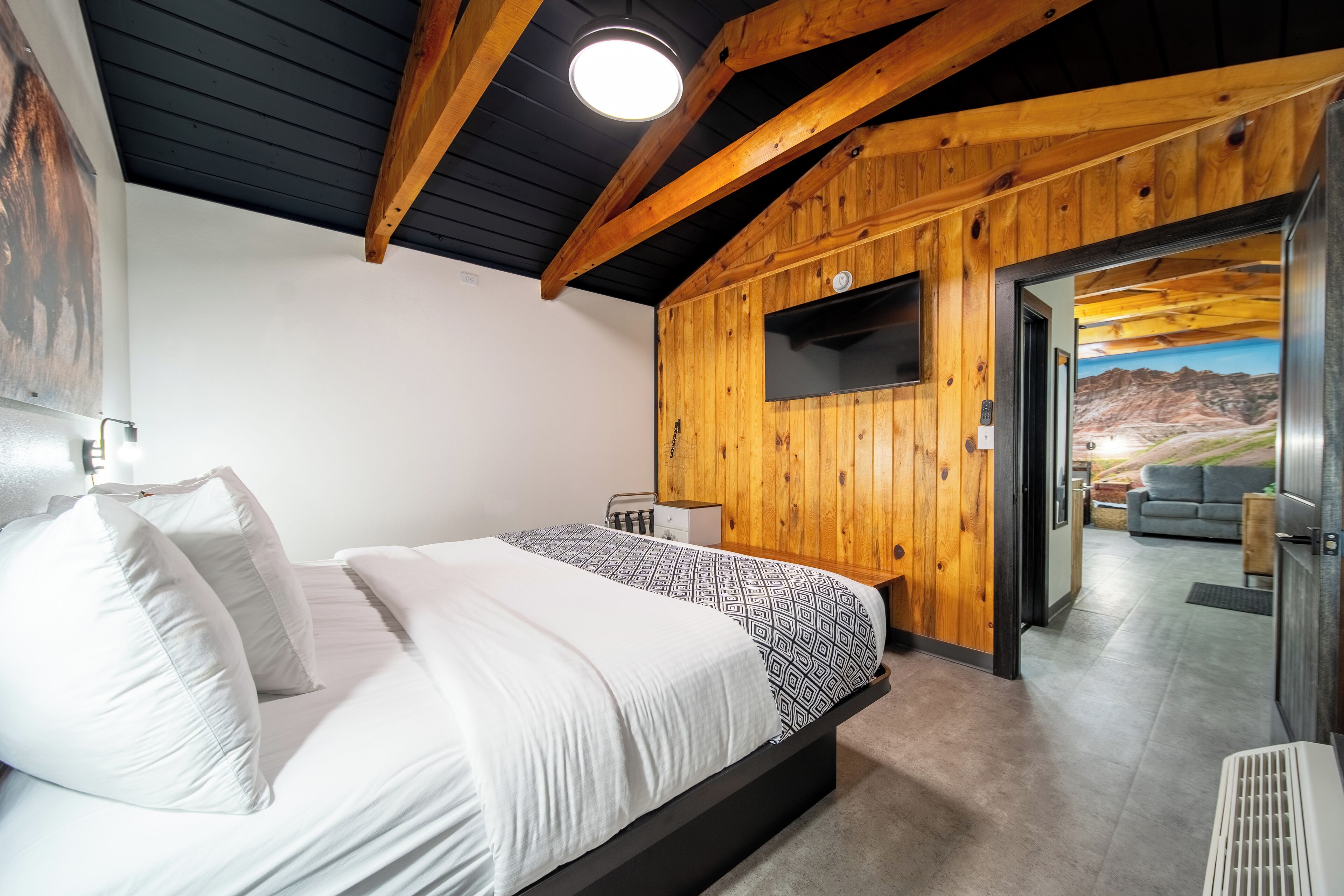 A bedroom with a wooden ceiling, a bed with white and patterned bedding, a TV on the wall, and a door leading to a living area with a view of a mountainous landscape.