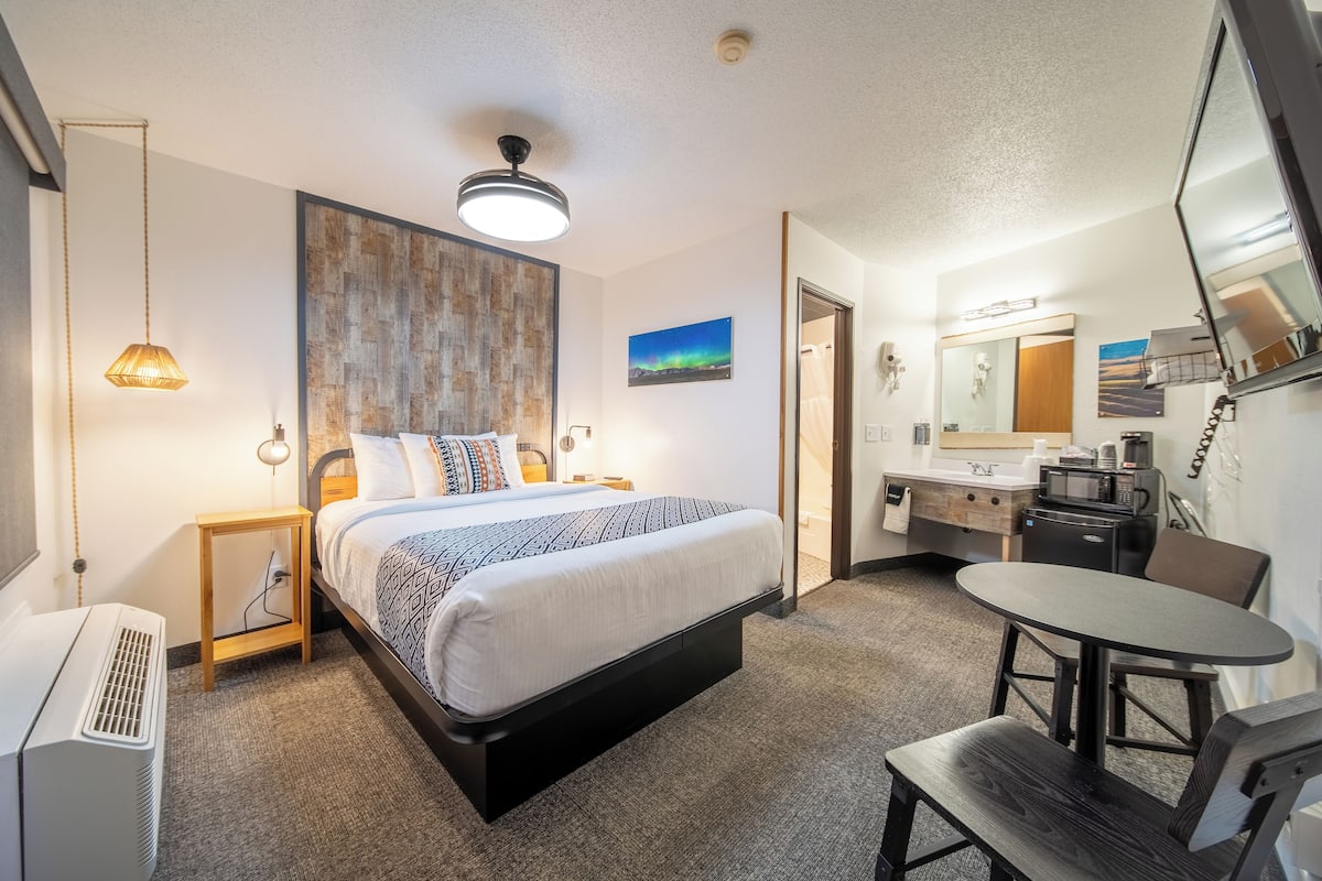 A hotel room with a bed, bedside tables, a desk, a chair, a small table, a microwave, a sink, and a mirror.