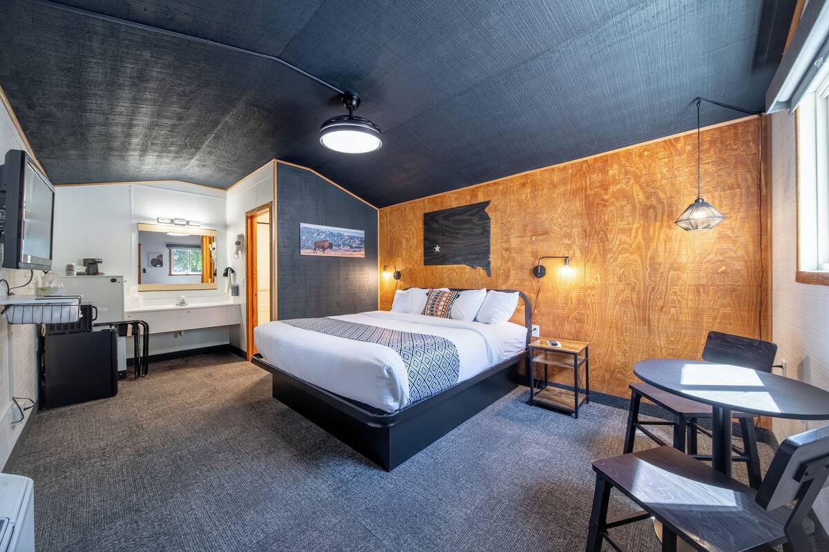 A modern hotel room with a large bed, a desk, and a chair. There is a bathroom with a sink and mirror. The room has a wooden accent wall and a ceiling light.