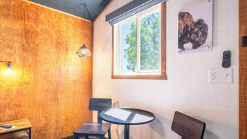 A cozy room with a wooden wall, a round table, two chairs, a bench, a window, and a wall-mounted light fixture.