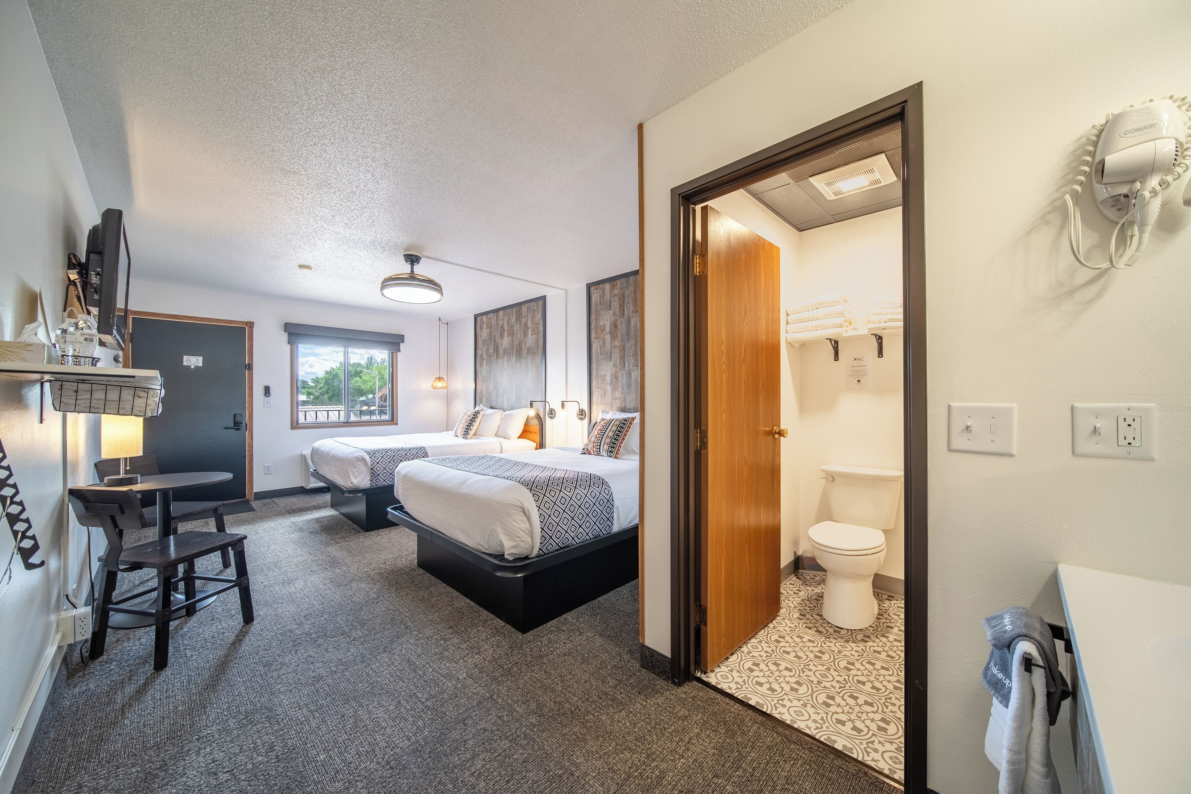A hotel room with two beds, a desk, a chair, a lamp, a hairdryer, and a bathroom with a toilet and sink.