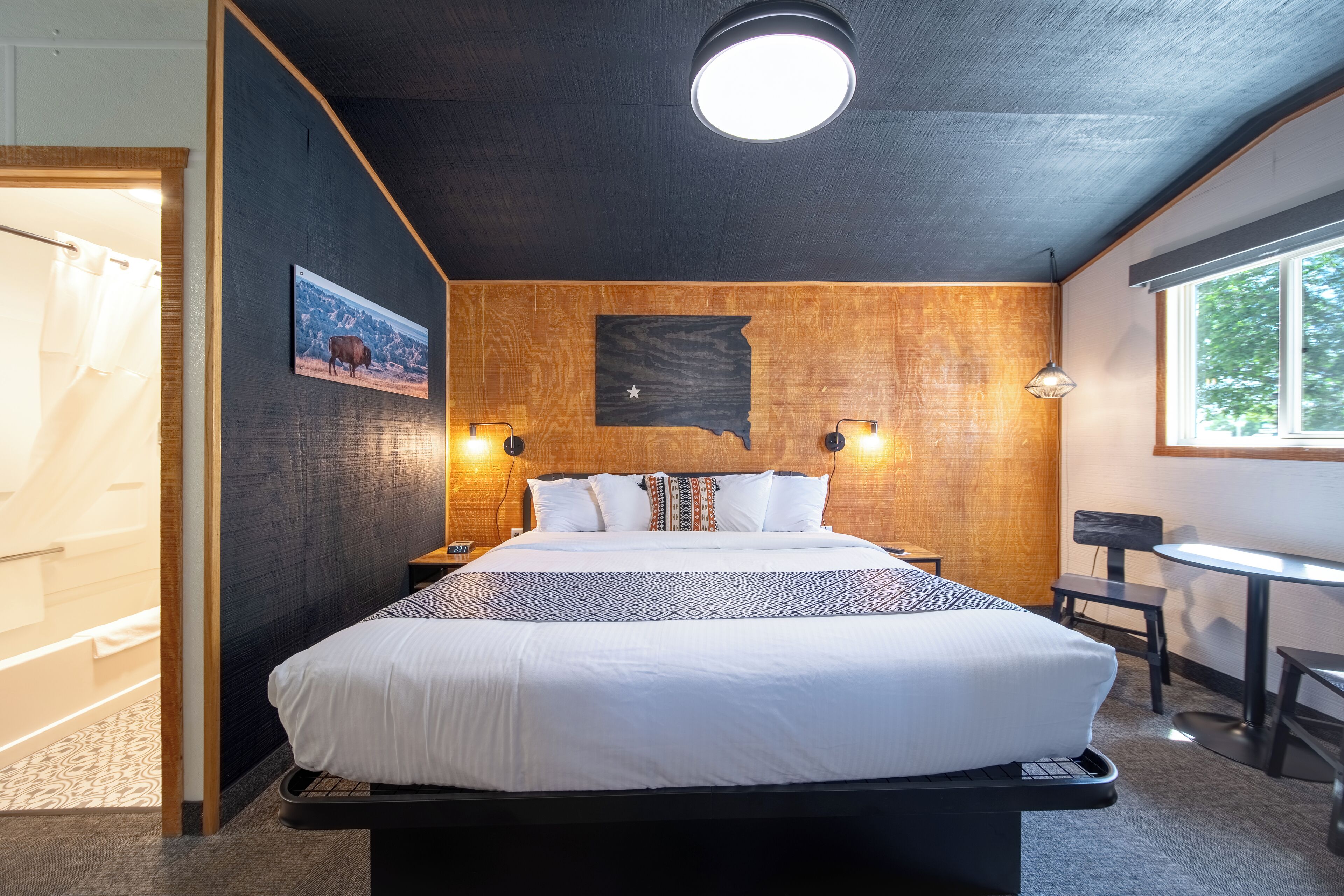 A bedroom with a large bed, wooden headboard, a wall-mounted map, two bedside lamps, and a window with a view of trees.
