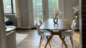 A dining area with a wooden table, white chairs, and a white rug. There is a window with curtains, a white chair with a decorative vase, and a plant on a side table. - Red Cabin Lake House with Amazing Views! (Vernon Township)