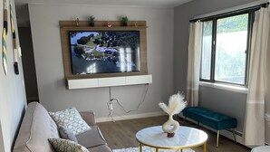 A modern living room with a sofa, a round coffee table, a TV mounted on the wall, and a window with curtains. - Red Cabin Lake House with Amazing Views! (Vernon Township)