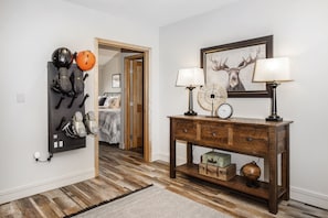 A room with a wooden floor, a wall-mounted rack with helmets, a wooden console table with lamps, and a framed deer picture.