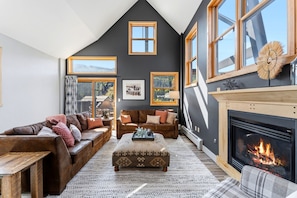 A cozy living room with a fireplace, leather sofas, a patterned ottoman, and a large window overlooking a scenic view.
