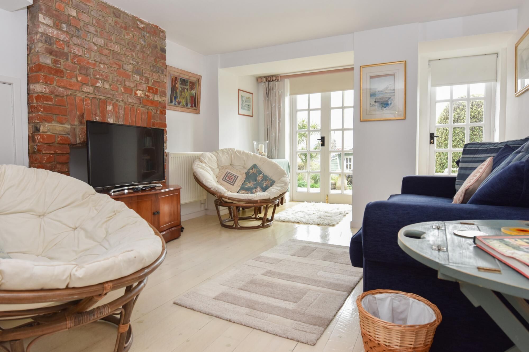 A living room with a brick fireplace, a television, a wicker chair, a blue sofa, and a large window. - Mulberry Cottage Coastal Holiday Home (Cowes)