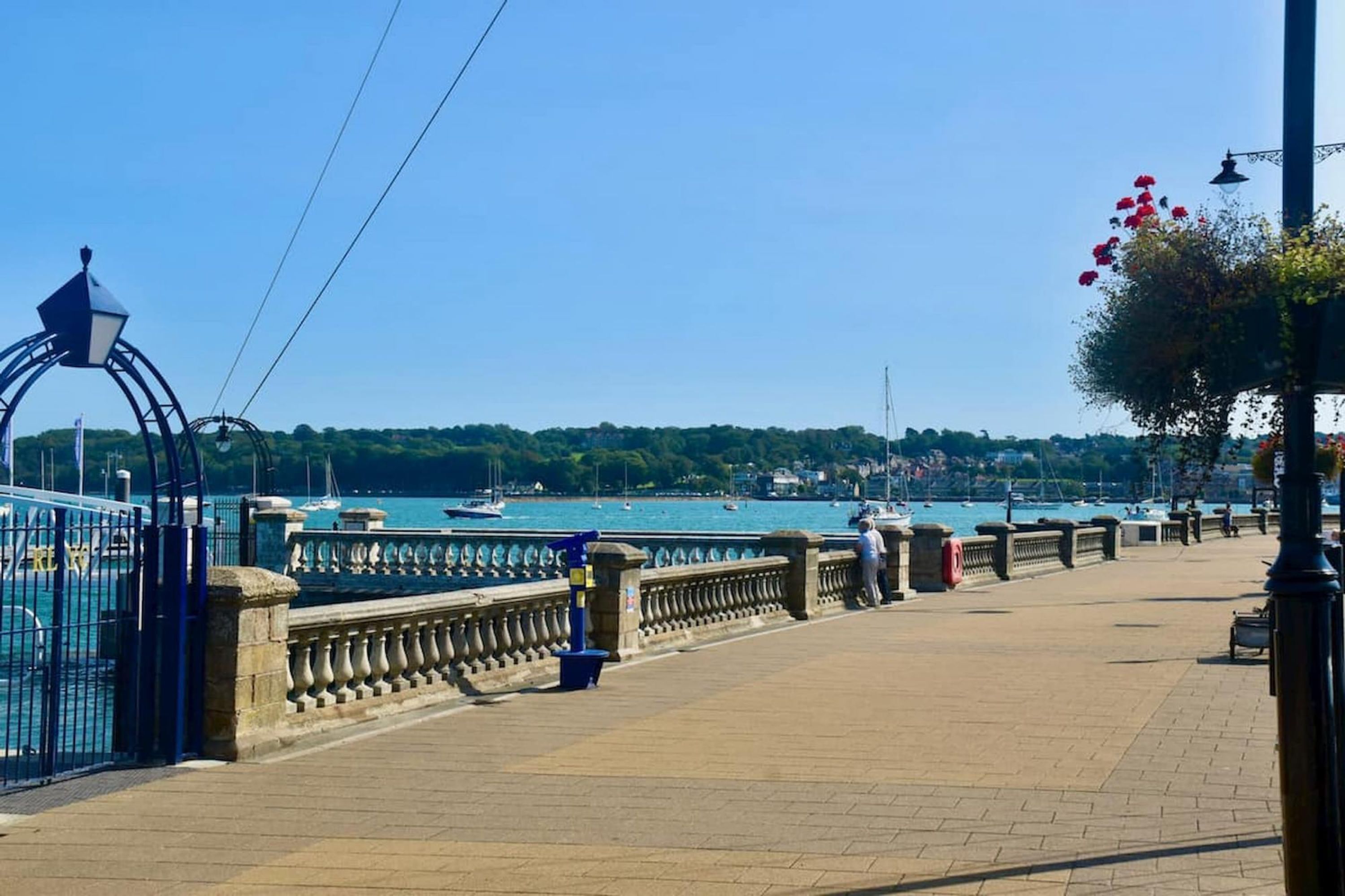 A waterfront promenade with a stone balustrade, a metal gate, and a row of street lamps.