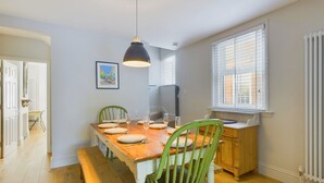 A dining area with a wooden table set for four, green chairs, a pendant light, and a window with blinds. - Blackberry Cottage Bembridge Accessible Coastal Holiday Home (Bembridge)