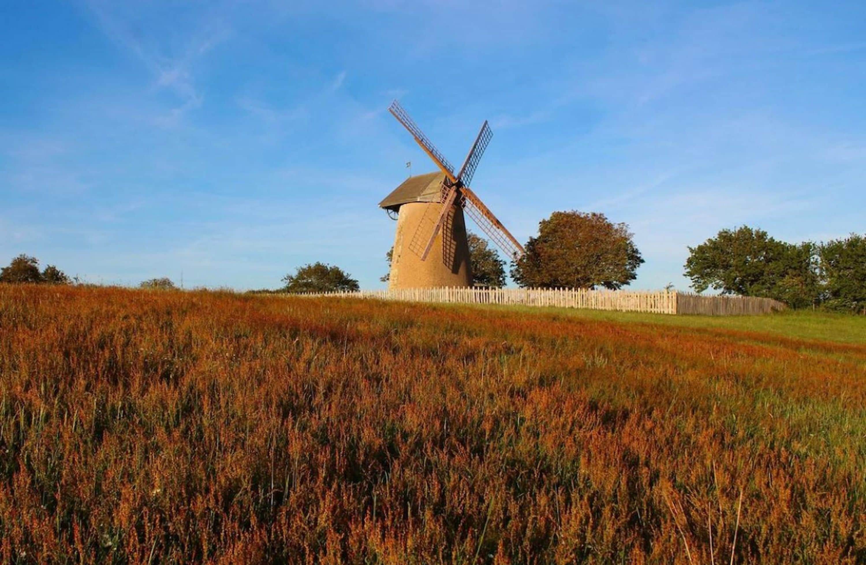 A windmill in a field with a wooden fence and trees in the background.