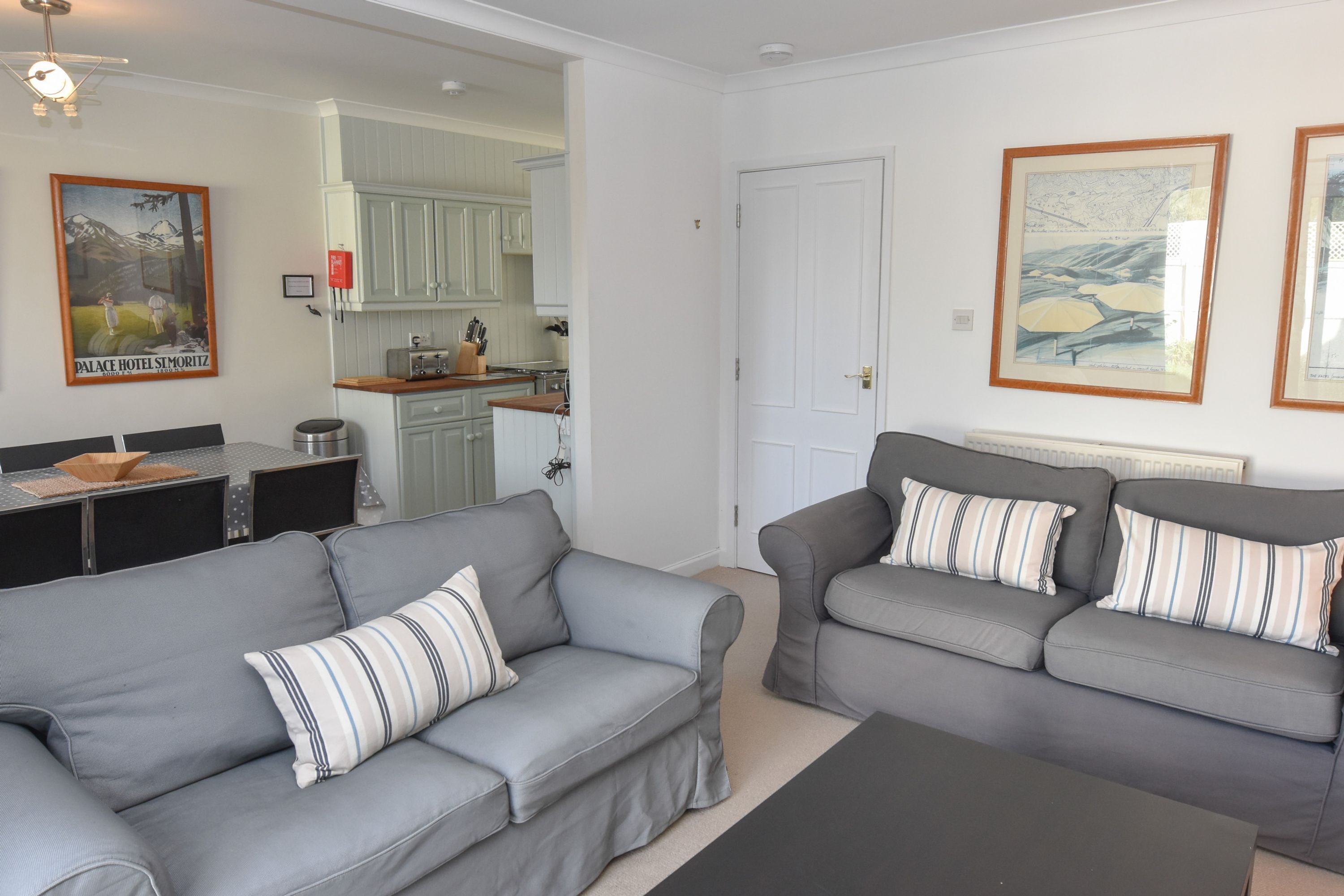 A living room with two grey sofas, a coffee table, and a dining area with a table and chairs. There is a kitchen visible in the background.