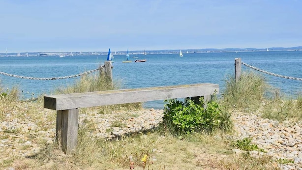 A wooden bench by a rocky shore with a view of the sea and distant cityscape. - Solent Landing No 12A - Beachfront (Bembridge)