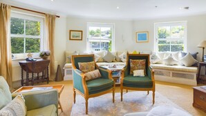 A spacious living room with two green armchairs, a sofa, and a coffee table. There are large windows with curtains, a radiator, and a lamp on a side table. - Solent Landing No 16 Beach Retreat (Bembridge)