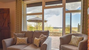 A cozy room with a sofa and armchair facing large windows, offering a view of a garden and hills.