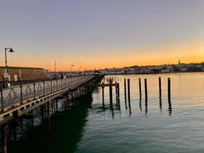 Eine Pier am Wasser mit einem Gehweg, im Hintergrund eine Stadtansicht und ruhiges Wasser.