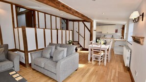 A modern living room with a grey sofa, wooden beams, a dining area, and a kitchen in the background. - The Cote Little Upton Farm (Ryde)