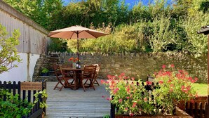 A wooden deck with a table and chairs under an umbrella, surrounded by a stone wall and lush greenery. - The Cote Little Upton Farm (Ryde)