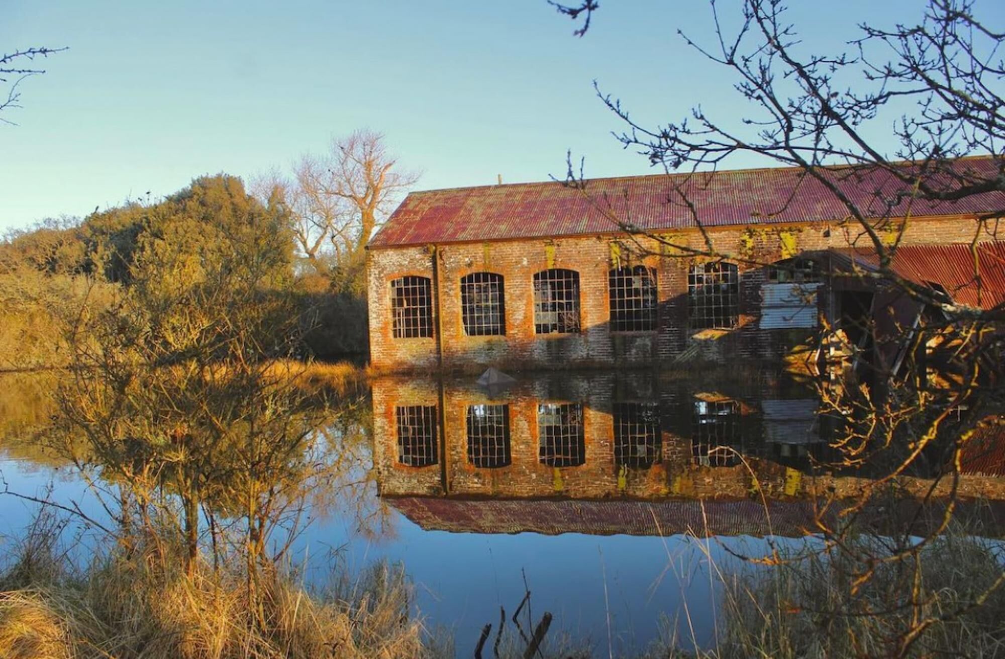A large, historic brick building with a red roof, reflecting in a calm body of water.