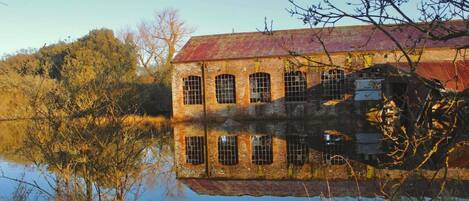 A large, historic brick building with a red roof, reflecting in a calm body of water.