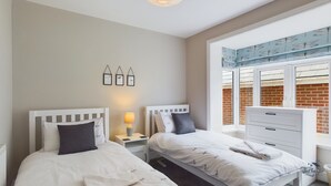 A bedroom with two beds, a nightstand with a lamp, a dresser, and a window with a view of a brick wall. - Coastal East View in St Helens (Ryde)
