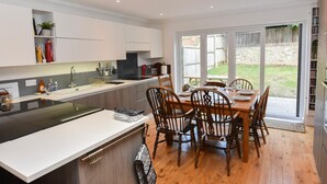 A modern kitchen with a central island, wooden dining table, and chairs. There is a large window providing a view of the backyard. - Queens Mews a Coastal Holiday Home (Ryde)