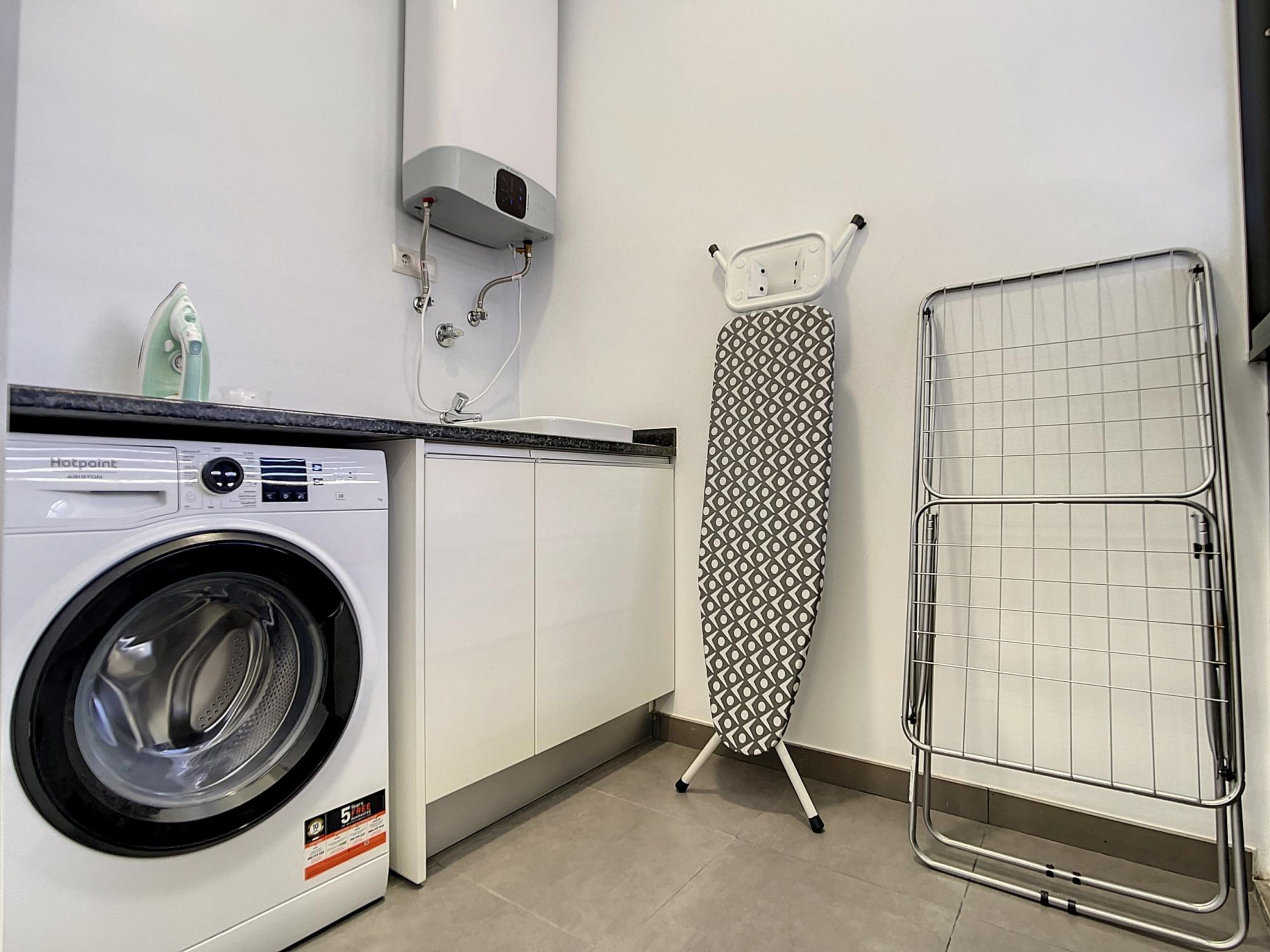 Un espace buanderie équipé d’un lave-linge à chargement frontal, d’une planche à repasser et d’un étendoir.