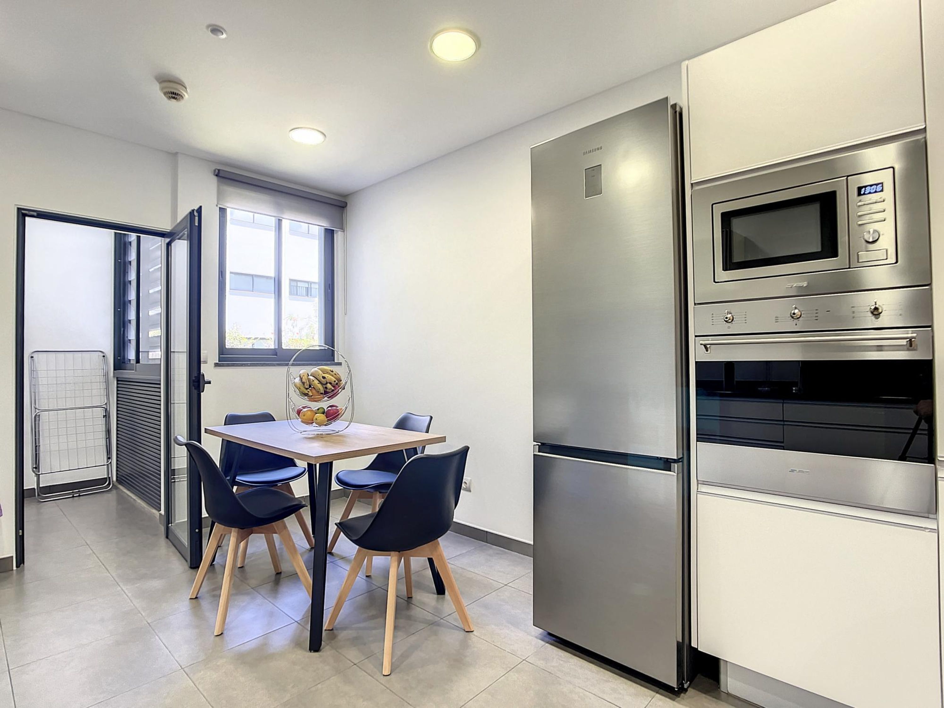 Una cocina moderna con un refrigerador, un microondas y un horno de acero inoxidable. Hay una mesa de comedor con sillas y una cesta de frutas encima.