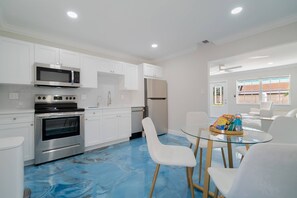 Een moderne keuken met witte kasten, roestvrijstalen apparatuur en een blauwe vloer. Er is een eethoek met witte stoelen en een glazen tafel.