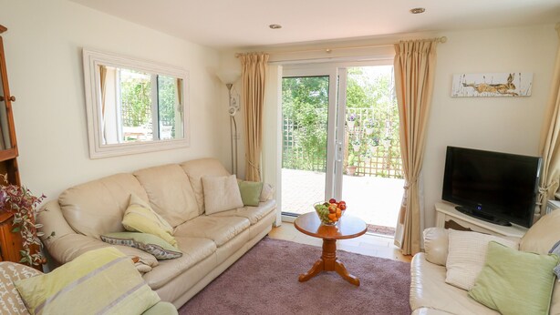 Ein Wohnzimmer mit beiger Couch, weißer Couch, Fernseher auf einem Schrank, einem Glastisch und Blick auf einen Garten durch eine Schiebetür.