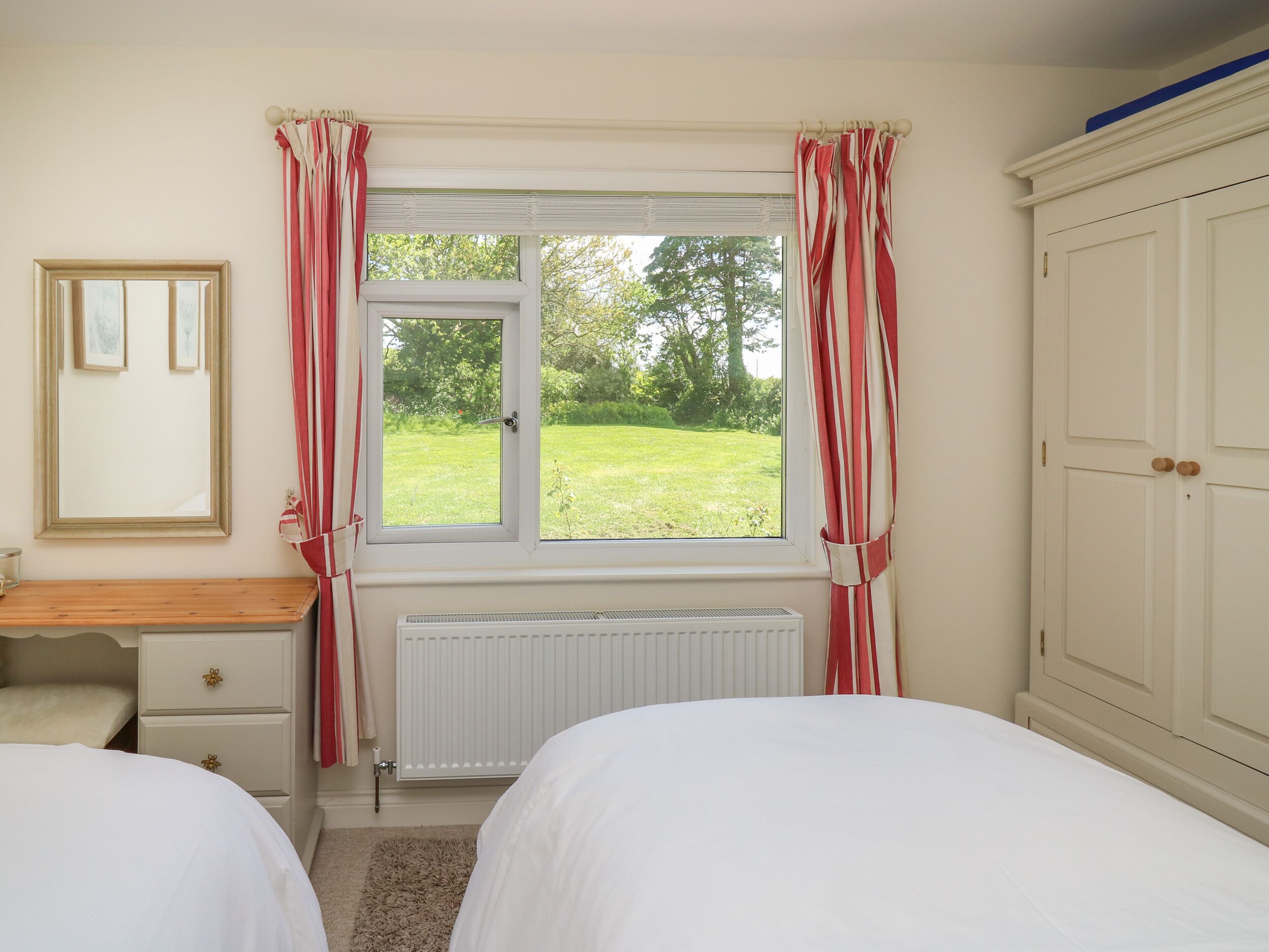 Ein Schlafzimmer mit zwei Betten, einem Fenster mit Vorhängen, einem Heizkörper, einem Holztisch, einem Spiegel und einem Kleiderschrank.