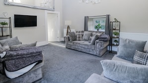 A living room with a grey sofa set, a large mirror, a flat-screen TV, and a 'HOME is where your heart is' sign.
