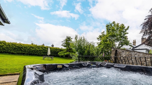 A hot tub in a garden with a picnic table and umbrella, surrounded by greenery and stone walls.