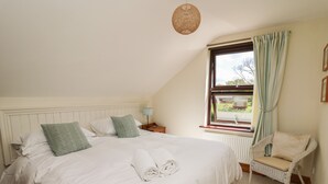Ein ordentlich bezogenes Bett mit Kissen, ein Nachttisch mit Lampe, ein Korbstuhl und Blick ins Freie durch ein Fenster mit Vorhängen.