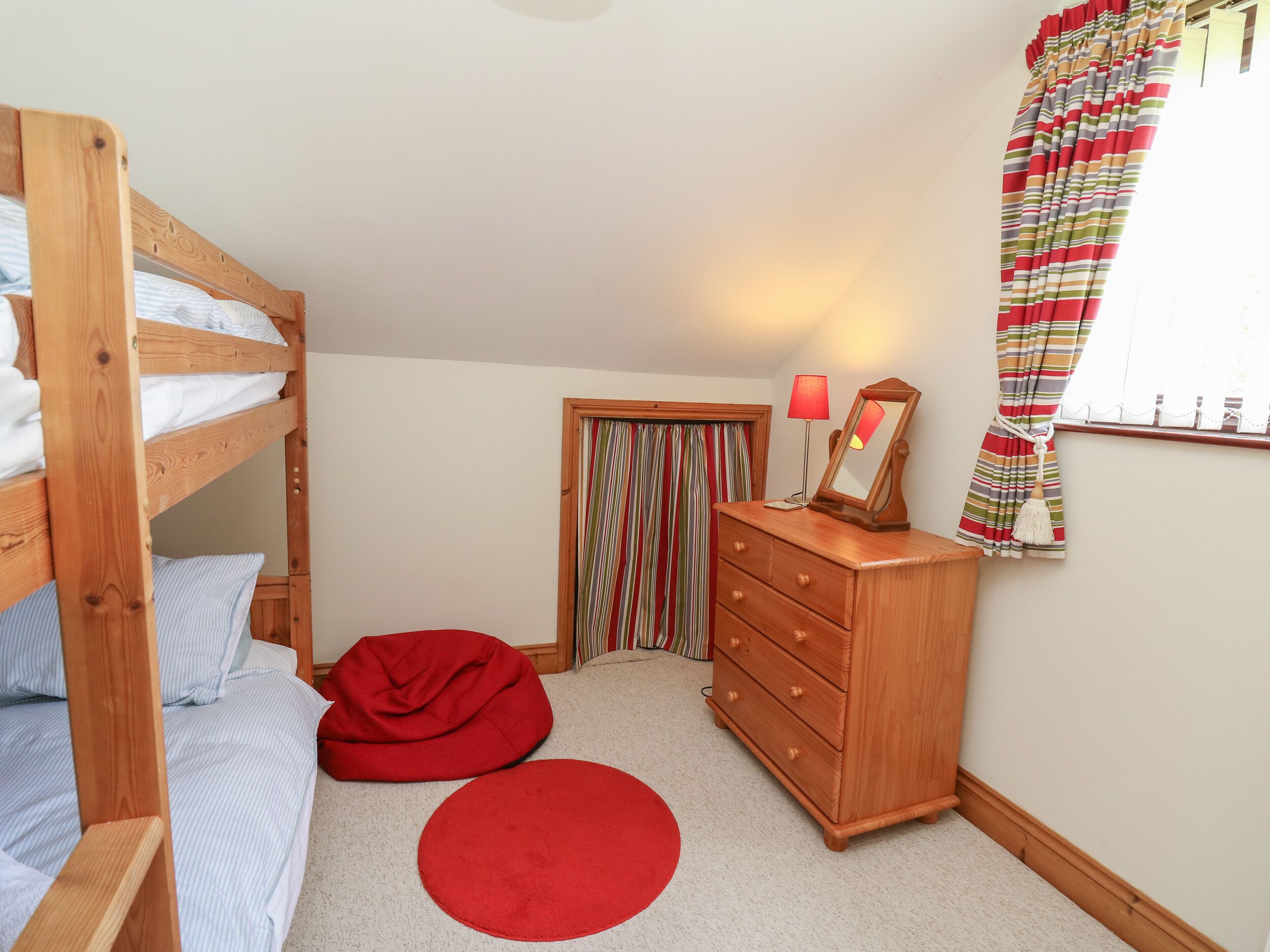 Een slaapkamer met een stapelbed, een dressoir, een spiegel, een lamp, een rode relaxstoel en een gestreept gordijn.