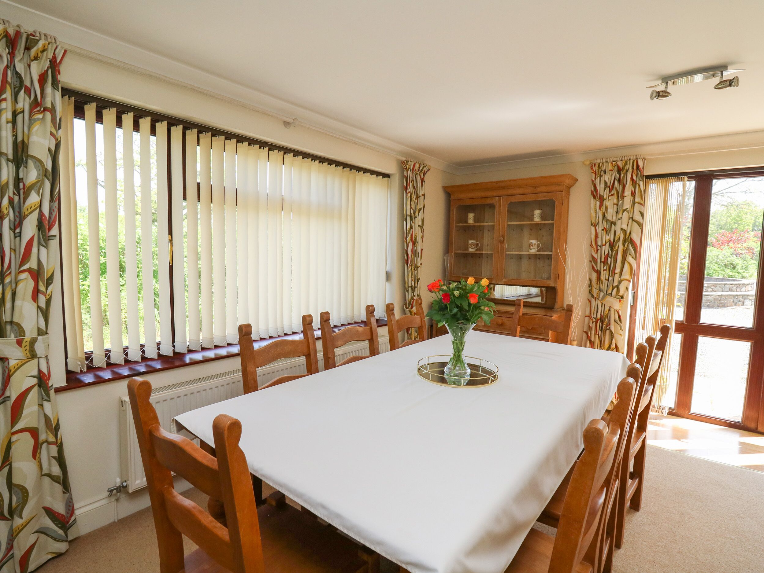A dining room with a large table, wooden chairs, a vase with flowers, and a cabinet with glass doors.