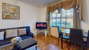 Ett vardagsrum med en soffa, ett soffbord med en vas, en tv som visar ”NETFLIX”, ett matbord med stolar och ett fönster med gardiner.