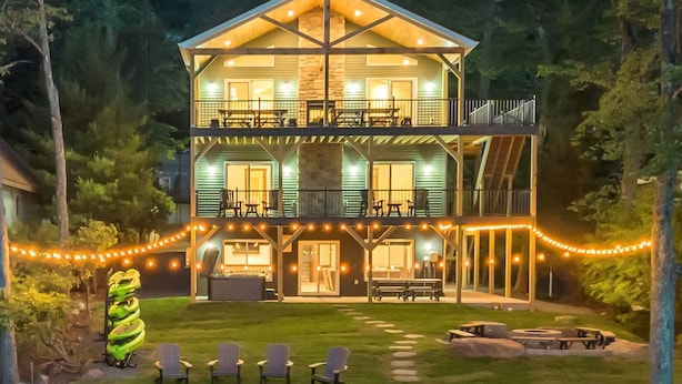 A two-story wooden cabin with a covered porch, outdoor seating, and a pond. - Lake Front Livin by PMR, 8BR Lakefront, Private Beach & Dock, Sleeps 26 (Lake Harmony)